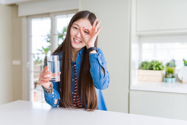 Mooie Aziatische vrouw drinken van een glaasje vers water met een blij gezicht lachend doen ok teken met de hand op oog kijken door de vingers - Foto, afbeelding