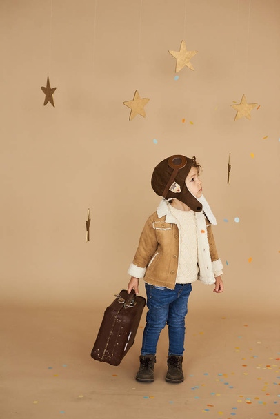 concept of dreams and travels. pilot aviator child with a toy airplane and suitcase plays in a beige background - Foto, Imagem