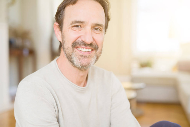 Handsome middle age man sitting on the floor smiling at the camera at home - 写真・画像