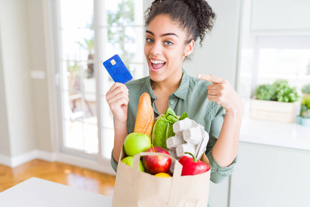 Jeune fille afro-américaine tenant sac en papier d'épicerie et carte de crédit comme paiement très heureux pointant avec la main et le doigt
 - Photo, image