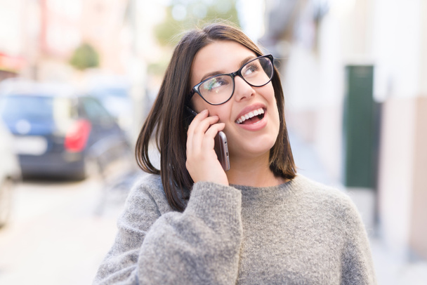 Jovem mulher bonita usando óculos sorrindo confiante falando ao telefone e andando na rua na cidade
 - Foto, Imagem