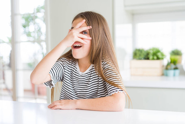 Beautiful young girl kid wearing stripes t-shirt peeking in shock covering face and eyes with hand, looking through fingers with embarrassed expression. - Фото, изображение