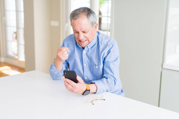 Handsome senior man using smartphone sending message annoyed and frustrated shouting with anger, crazy and yelling with raised hand, anger concept - Fotoğraf, Görsel