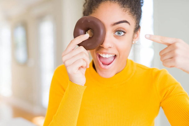 Jovem afro-americana comendo doce donut de chocolate muito feliz apontando com a mão e o dedo
 - Foto, Imagem