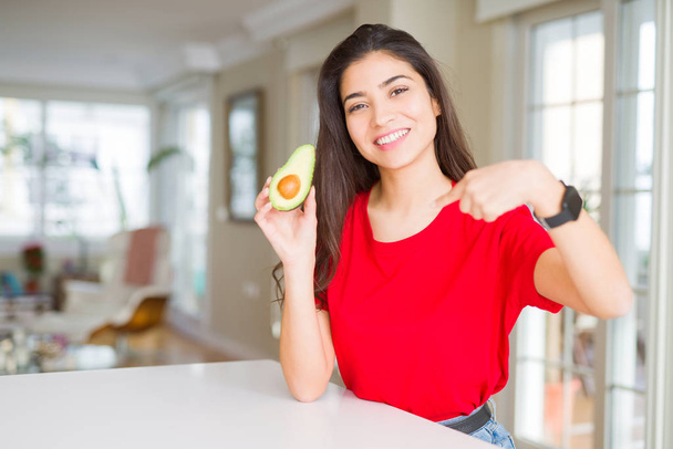 Young woman eating healthy avocado with surprise face pointing finger to himself - Photo, Image