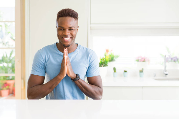 Przystojny mężczyzna african american noszenie t-shirt na co dzień w domu modlitwy rękoma razem z prośbą o przebaczenie uśmiechający się pewnie. - Zdjęcie, obraz