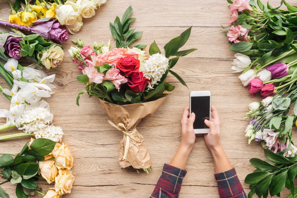 Частичный вид флориста с помощью смартфона с чистым экраном рядом с цветами на деревянной поверхности
 - Фото, изображение