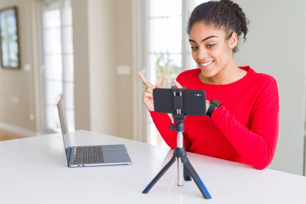 Молодая африканская американка делает видеозвонок с помощью камеры смартфона улыбаясь и глядя на камеру указывая двумя руками и пальцами в сторону
. - Фото, изображение