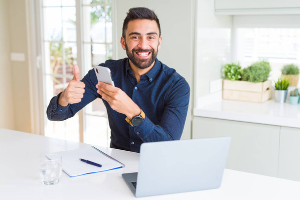 Knappe Spaanse zakenman met smartphone en laptop op het kantoor gelukkig grote glimlach doen ok teken, duim omhoog met vingers, uitstekende teken - Foto, afbeelding