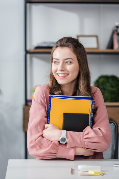 attraktive Studentin im rosa Hemd hält lächelnd Notizbücher in der Hand - Foto, Bild