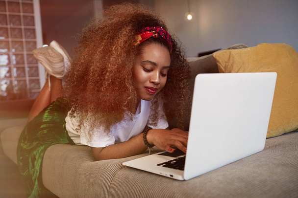 Kaunis amerikkalainen afrikkalainen nainen kirjoittamassa kannettavalla, tekstailemassa ystäville sosiaalisten verkostojen kautta. Opiskelija tyttö selailee Internetiä, käyttäen wi-fi, istuu sohvalla kotona
. - Valokuva, kuva