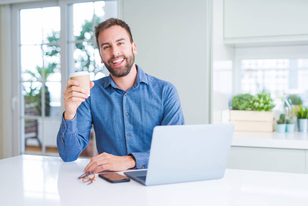 schöner Mann, der mit einem Laptop arbeitet und eine Tasse Kaffee trinkt, mit einem glücklichen Gesicht im Stehen und einem selbstbewussten Lächeln, das Zähne zeigt - Foto, Bild
