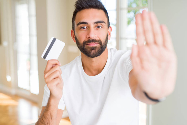 Красивый латиноамериканец держит кредитку с открытой рукой делает знак стоп с серьезным и уверенным выражением, защитный жест
 - Фото, изображение