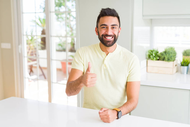 Camiseta amarilla casual de hombre hispano guapo en casa haciendo un gesto feliz con la mano. Aprobar la expresión mirando a la cámara con éxito
. - Foto, imagen