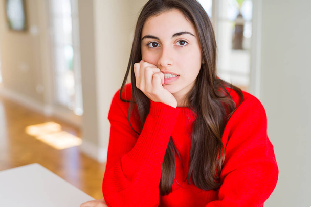Красива молода жінка в повсякденному червоному светрі виглядає напруженою і нервовою з руками на роті кусає цвяхи. Проблема тривоги
. - Фото, зображення