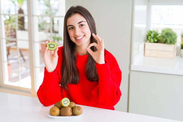 Beautiful young woman eating half fresh green kiwi doing ok sign with fingers, excellent symbol - Фото, зображення