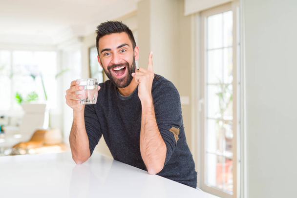 Hombre hispano guapo bebiendo un vaso de agua fresca sorprendido con una idea o pregunta apuntando con el dedo con la cara feliz, número uno
 - Foto, imagen