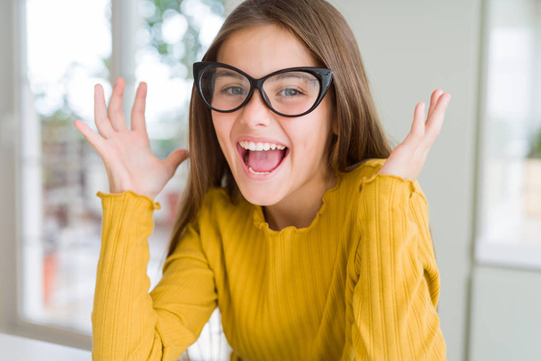 Beautiful young girl kid wearing glasses celebrating crazy and amazed for success with arms raised and open eyes screaming excited. Winner concept - Zdjęcie, obraz