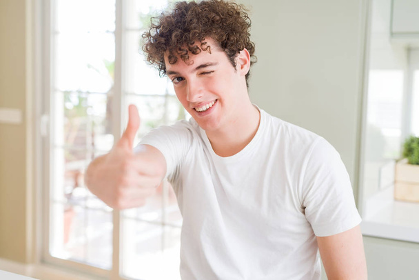 Joven hombre guapo usando una camiseta blanca haciendo un gesto feliz con la mano. Aprobación de la expresión mirando a la cámara mostrando éxito
. - Foto, imagen