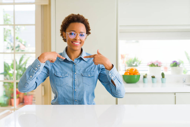 Jeune belle femme afro-américaine portant des lunettes regardant confiant avec le sourire sur le visage, se montrant du doigt fier et heureux
. - Photo, image
