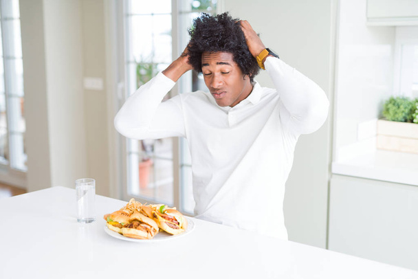Afrikkalainen amerikkalainen nälkäinen mies syö hampurilaisia lounaaksi kärsii päänsärystä epätoivoinen ja stressaantunut, koska kipu ja migreeni. Kädet pään päälle.
. - Valokuva, kuva