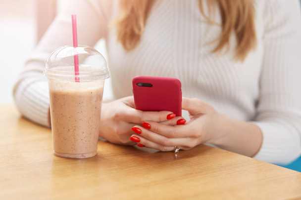 Mobile smart phone in blonde female hands with bright red manicure, puts arms on table near cocktail, finds information in gadget or types message for her friend, uses wireless Internet in cafe. - Фото, изображение
