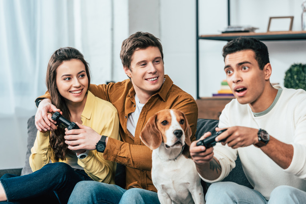 Три багатоетнічні друзі з бігль-собакою тримають джойстики і грають у відеоігри
 - Фото, зображення