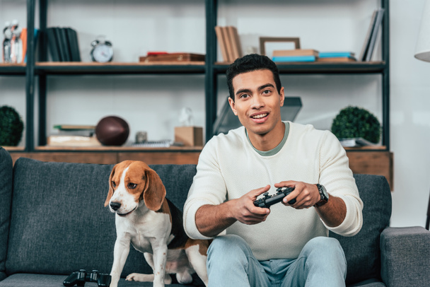 Jonge man met hond zittend op sofa en holding joystick - Foto, afbeelding