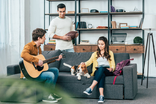 Three multicultural friends with beagle dog playing guitar in living room - Foto, immagini