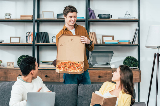 Hymyilevä nuori mies oranssissa paidassa näyttää pizzaa ystäville olohuoneessa
 - Valokuva, kuva