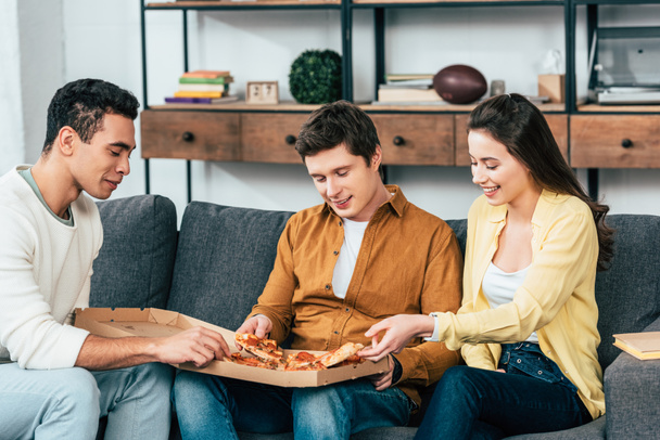 Three joyful multiethnic friends sitting on sofa and eating pizza together - Foto, afbeelding