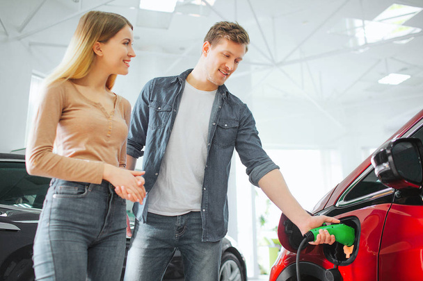 Young smiling couple buying first electric car in the showroom. Man charging modern eco-friendly vehicle with the power cable supply plugged in. The future of the automobile - Foto, imagen