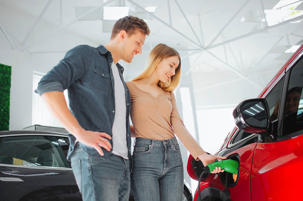 Young smiling couple buying first electric car in the showroom. Woman charging modern eco-friendly vehicle with the power cable supply plugged in - Photo, Image