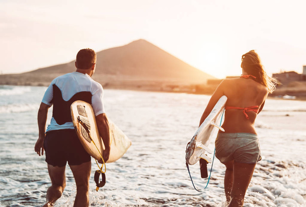 Happy surfer couple running with surfboards along the sea shore - Sporty people having fun going to surf together at sunset - Extreme surfing sport and youth relationship lifestyle concept - Photo, Image