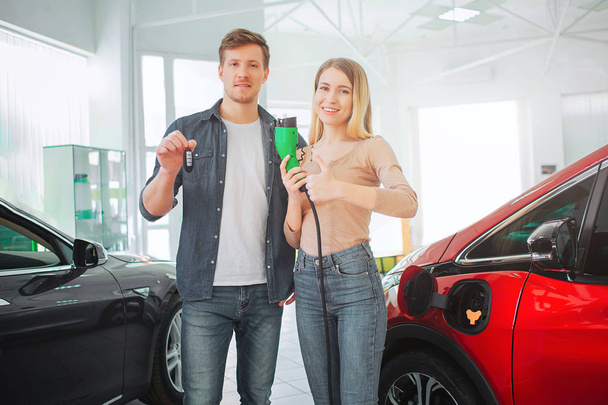 Young smiling family buying first electric car in the showroom. Environmental protection. Attractive man and woman holding car key and electric car charging plug. Green electric car sale concept - Photo, Image