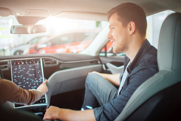 Young couple buying first electric car in the showroom. Attractive glad man looking at his girlfriend while she chooising a way on electronic dashboard in modern electric hybrid vehicle before test - Foto, Imagen