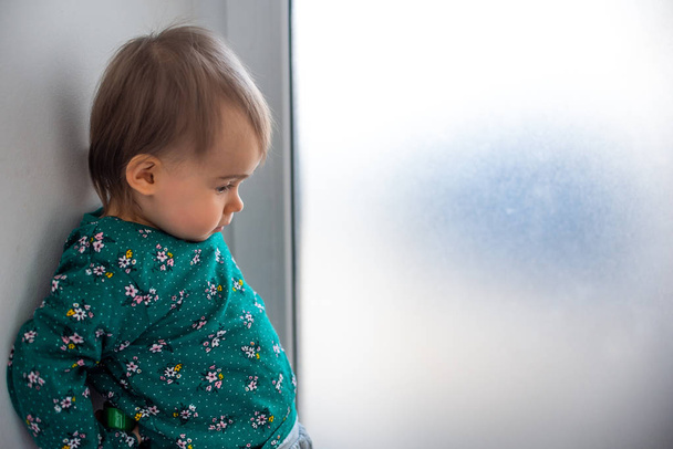 Cute caucasian one year old baby girl in green shirt standing with back against wall next to bright window. - Photo, Image
