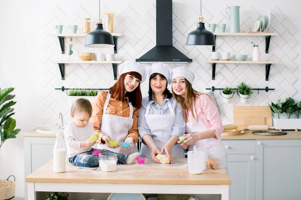 Happy family in the kitchen. Young woman and her sister, middle aged woman and little cute daughter cooking cupcakes for Mothers day, casual lifestyle photo series in real life interior - Foto, Imagen