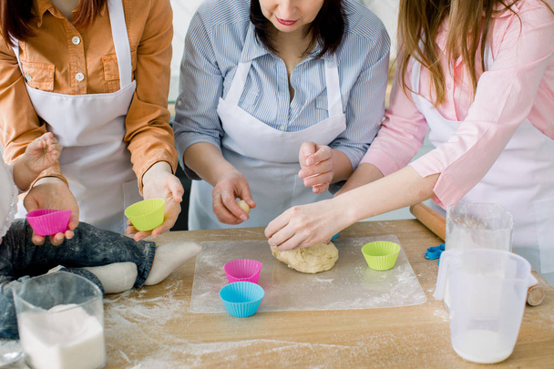 Happy women in white aprons baking together. Close-up photo of women hands and little baby preparing dough for baking muffins. Family, cooking, baking and people concept - Photo, Image