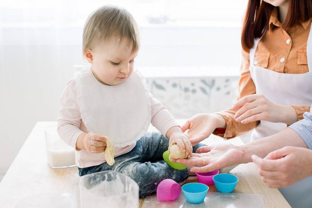 Close-up portrait of cute little baby girl sitting on the dinner table and playing with dough for baking muffins in coloured forms. Mother and grandmother working with dough. Mothers Day concept - Photo, Image