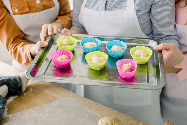 Preparing muffins for baking. Dough divided into silicone colourful cups lying on metal baking tray. Close-up of women hands holding muffin baking tray. - Photo, Image
