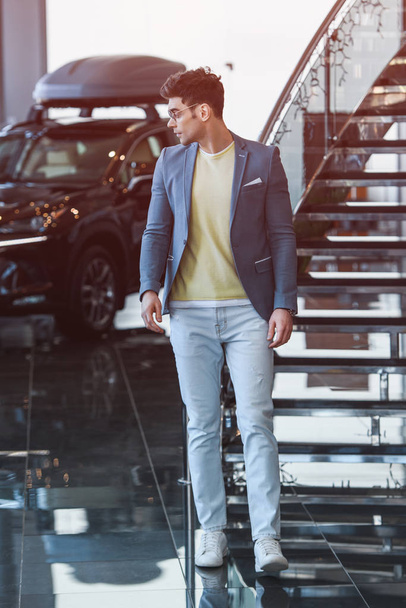 guapo hombre elegante en gafas de pie en sala de exposición de coches
  - Foto, Imagen