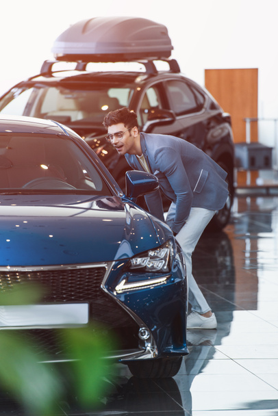 cheerful stylish man in glasses standing near automobile in car showroom  - Fotó, kép