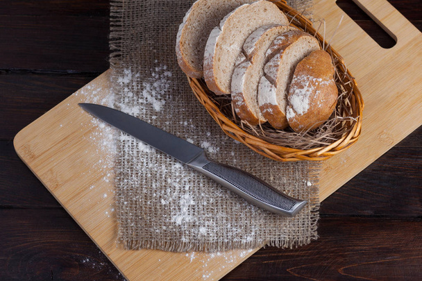 Свежий коричневый хлеб с нарезанными кусочками на дубовой доске с разбросанной мукой на деревянном фоне из льняной ткани со стальным ножом. Вид сверху
 - Фото, изображение