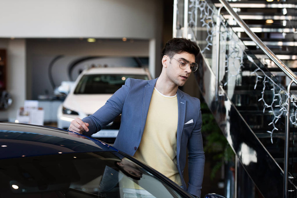 gutaussehende Kundin mit Brille steht neben Auto im Autohaus  - Foto, Bild