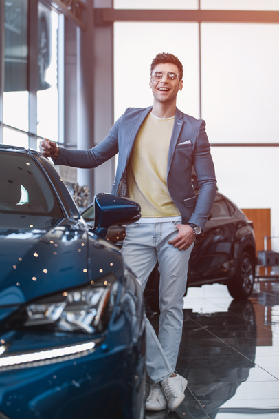 selective focus of cherful man in glasses standing near automobile with crossed legs in car showroom  - Photo, Image