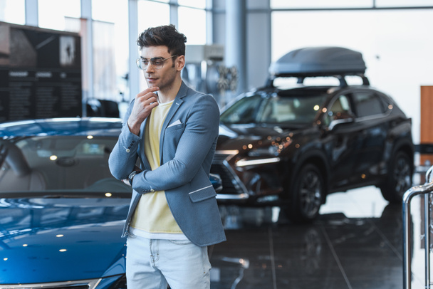 thoughtful man in glasses standing near automobiles in car showroom  - Fotoğraf, Görsel