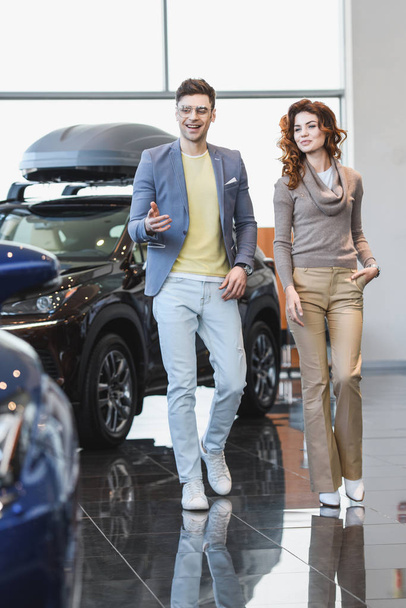 happy man in glasses pointing with finger at automobile near curly woman with hand in pocket  - Foto, Bild