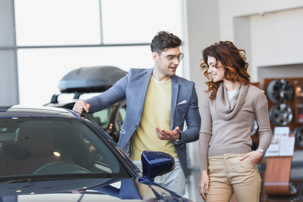 glücklicher Mann mit Brille zeigt mit dem Finger auf Automobil neben lockiger Frau mit Hand in der Tasche  - Foto, Bild