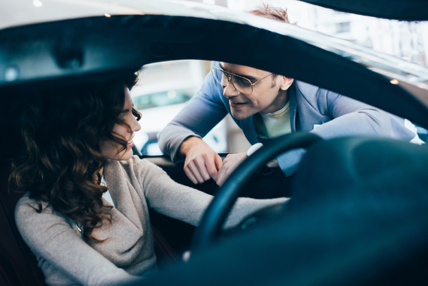 selective focus of curly woman sitting in automobile and holding steering wheel near cheerful man in glasses - 写真・画像